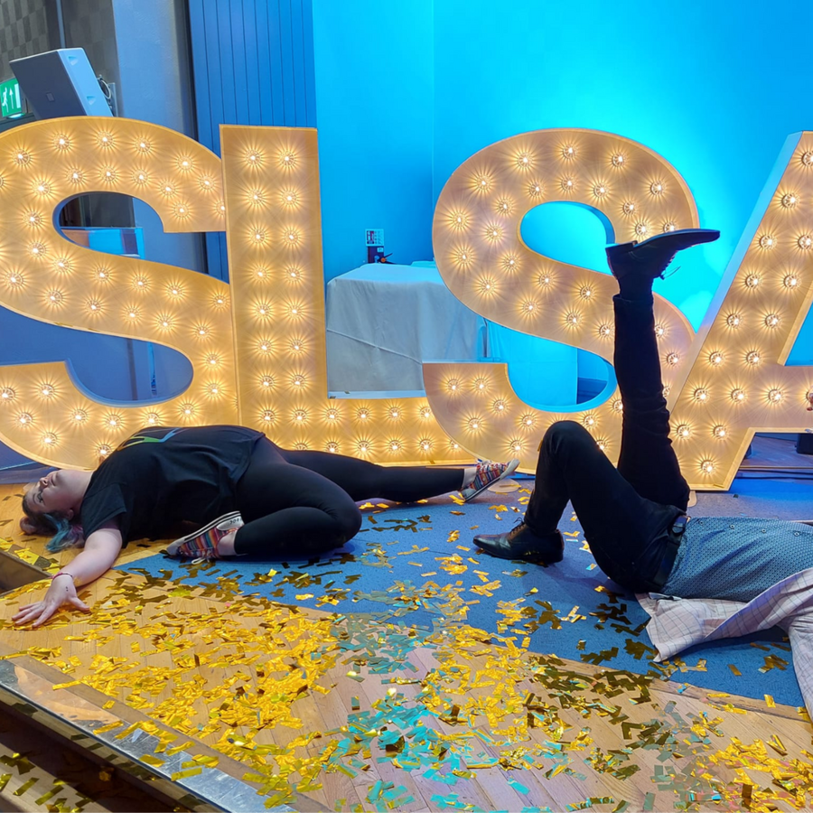 Two of our researchers, Rosie and James, having just death-dropped in front of the large "SLSA" letters on stage at an SLSA conference