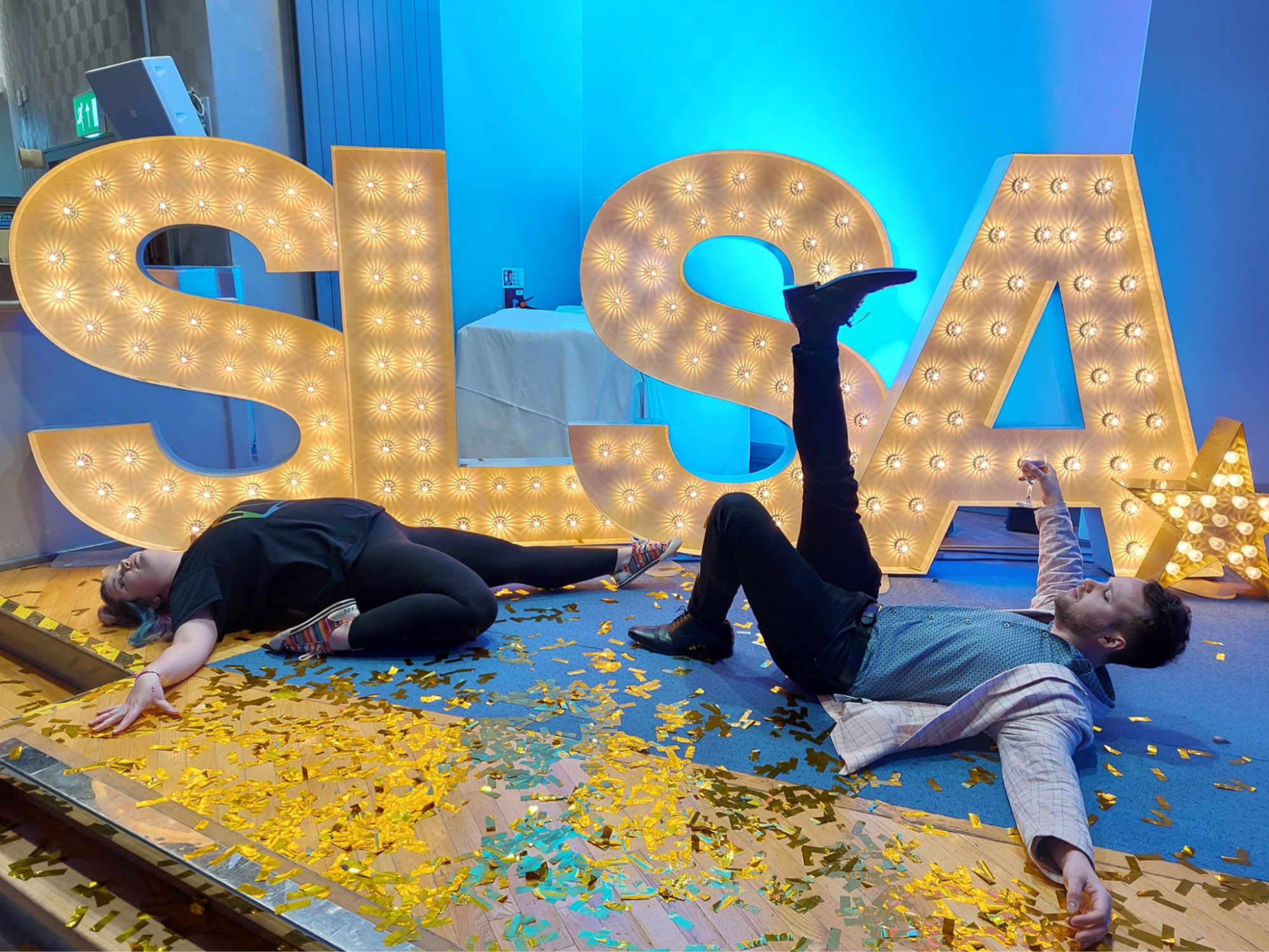 Rosie and James, having just death-dropped in front of the large "SLSA" letters on stage at an SLSA conference