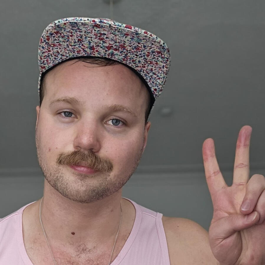James taking a pre-pride selfie: big gay moustasche and stubble, pink vest and short, pink polka dot hat, smiling and giving a peace sign